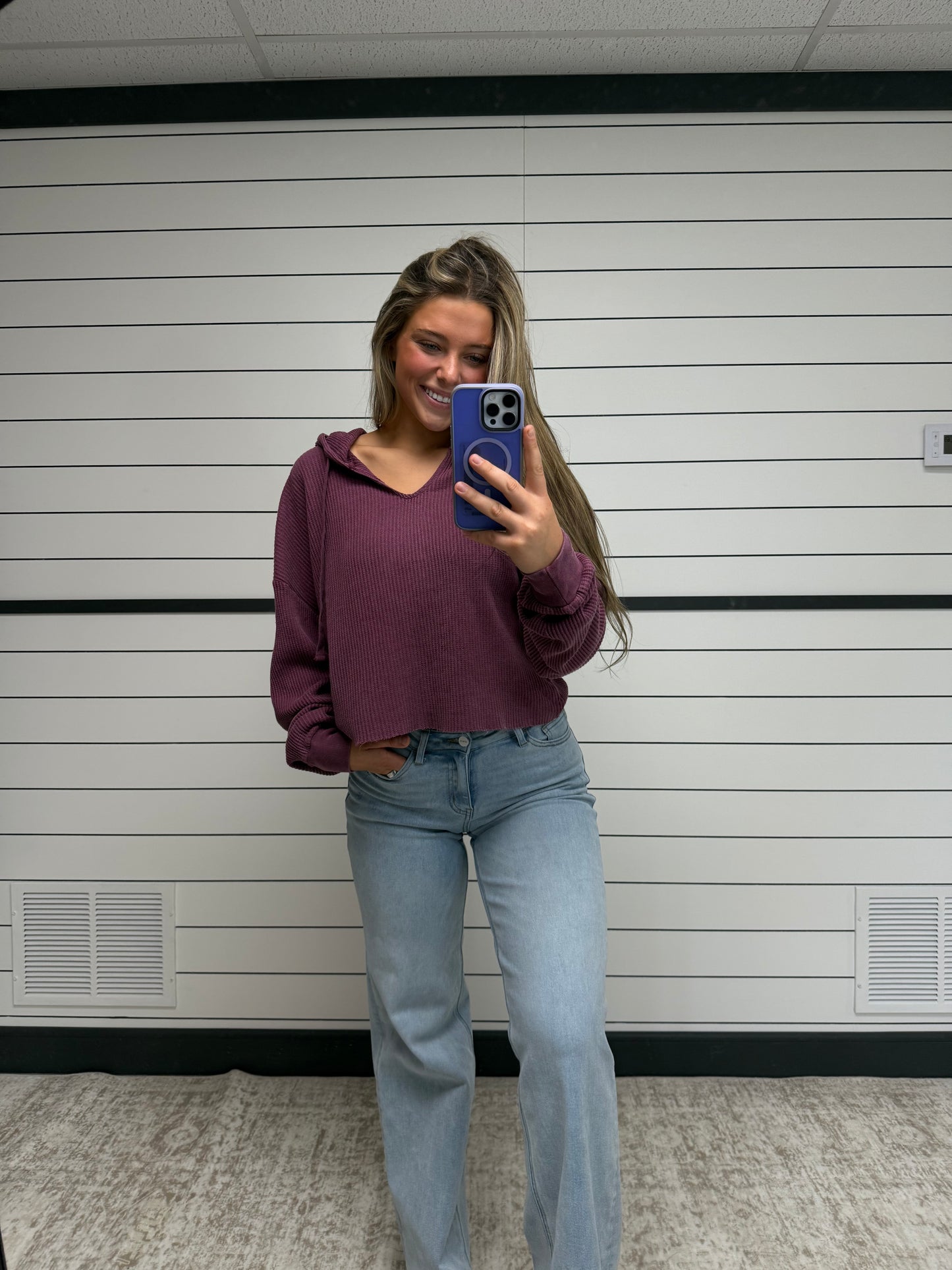Person taking a mirror selfie wearing a purple hoodie and blue jeans in a bathroom.
